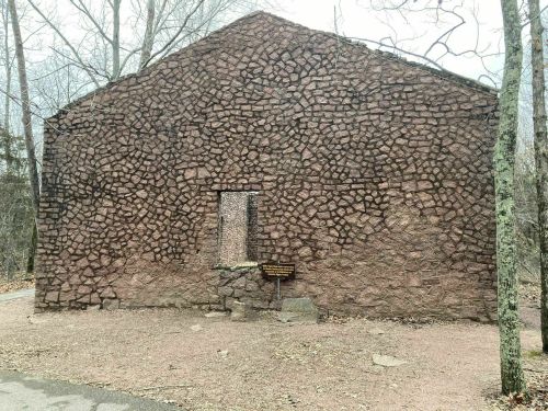 Stone building with a single window, surrounded by trees and a sign in front, set in a natural landscape.