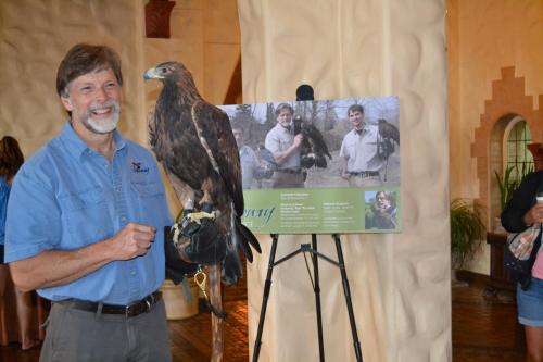 A man holds a hawk next to a display featuring his photo and information about his work with birds of prey.