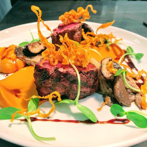 A beautifully plated dish featuring grilled steak, mushrooms, squash, and crispy garnishes on a white plate.