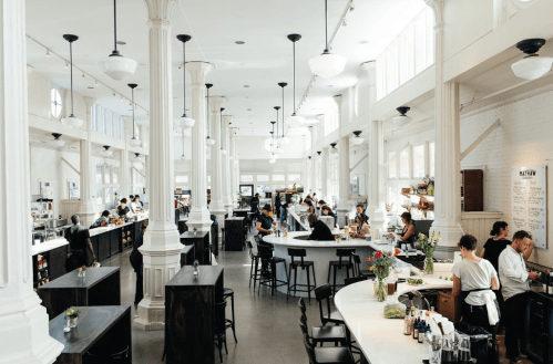 A spacious, bright café with tall columns, patrons at tables, and staff preparing food and drinks.