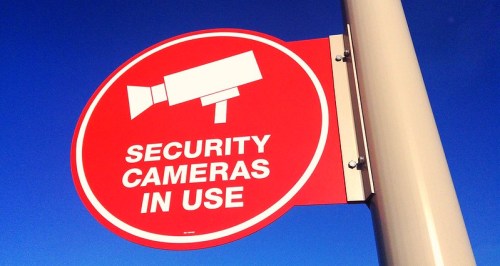 Red sign with a security camera icon and text: "SECURITY CAMERAS IN USE" against a blue sky.
