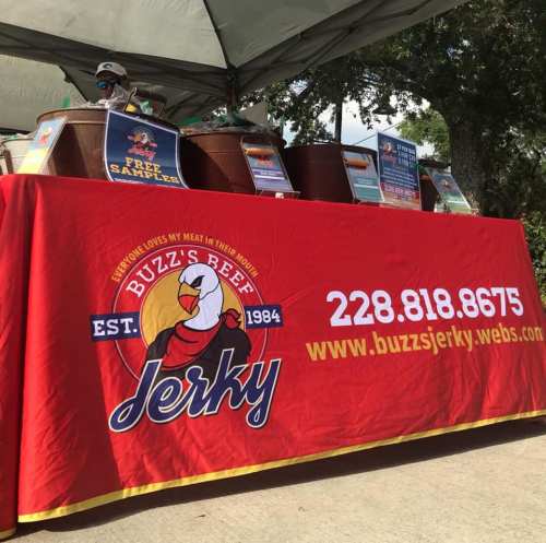 A colorful table display for Buzz's Beef Jerky, featuring samples and promotional materials under a tent.