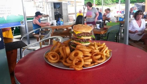 A towering burger with cheese and toppings sits on a plate surrounded by curly fries, in a busy outdoor dining area.