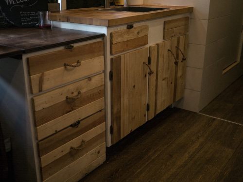 A rustic kitchen with wooden cabinets and drawers, featuring a sink and a dark countertop. Warm, natural tones throughout.