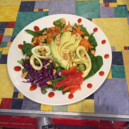 A colorful salad with spinach, red cabbage, carrots, avocado, bell pepper, and nuts, drizzled with sauce on a patterned table.
