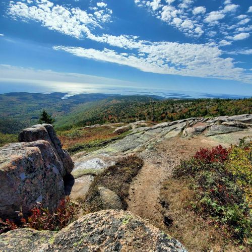 A scenic view from a rocky mountain top, showcasing lush green hills, colorful foliage, and a shimmering body of water.