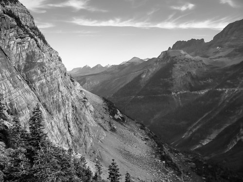 Black and white landscape of rugged mountains and steep cliffs, with a valley and scattered trees in the foreground.