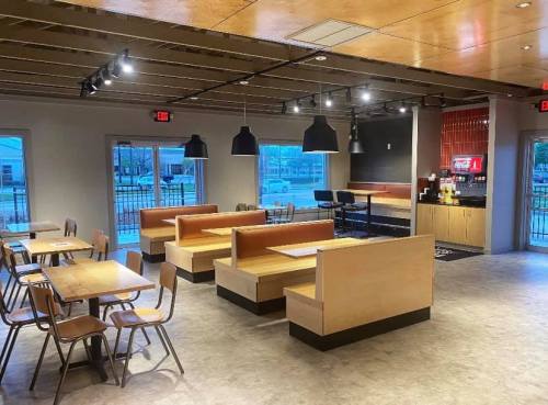 Modern restaurant interior with wooden tables, booths, and a beverage station, featuring large windows and warm lighting.