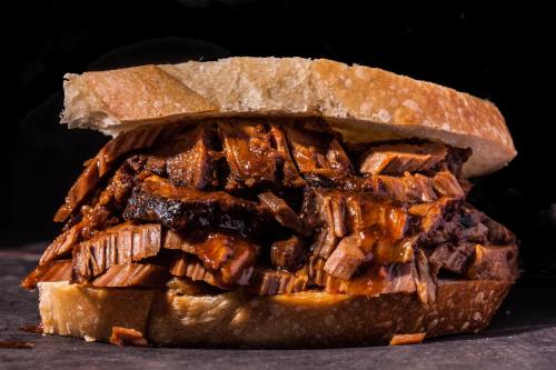 A close-up of a stacked brisket sandwich with barbecue sauce, served on thick slices of bread against a dark background.