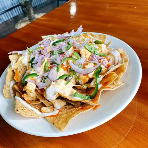 A plate of nachos topped with shredded chicken, onions, green peppers, and drizzled with sauce on a wooden table.