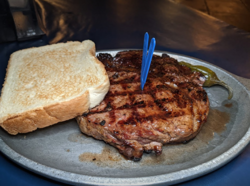 A grilled steak on a plate with a slice of toast and a blue toothpick, accompanied by a green pepper.