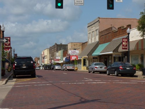 A quaint downtown street with brick pavement, shops, and traffic lights under a partly cloudy sky.