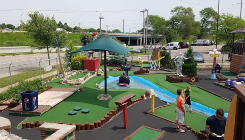 A colorful mini-golf course with a stream, sculptures, and players enjoying the game on a sunny day.