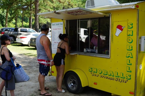 A bright yellow snoball food truck with customers ordering in a park setting. Trees and parked cars are in the background.