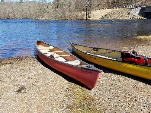Two canoes, one red and one yellow, are parked on the shore of a calm river surrounded by bare trees.