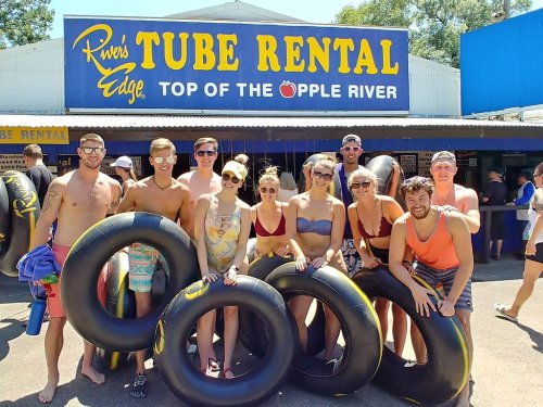 A group of young adults poses with inner tubes outside a tube rental shop on a sunny day.