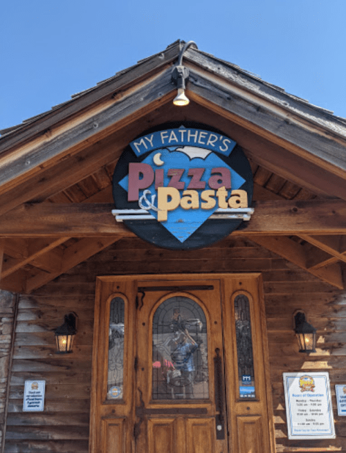 Sign for "My Father's Pizza & Pasta" on a wooden building with a rustic entrance and clear blue sky.