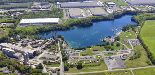 Aerial view of a lake surrounded by greenery, industrial buildings, and a parking area with numerous cars.