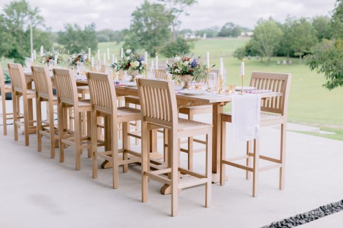A beautifully set outdoor dining table with wooden chairs, floral centerpieces, and candles, surrounded by greenery.