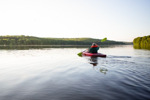 A person kayaking on a calm lake surrounded by lush green trees under a clear sky.
