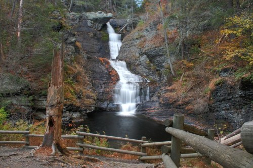 A serene waterfall cascades down rocky cliffs, surrounded by autumn foliage and a wooden fence in the foreground.