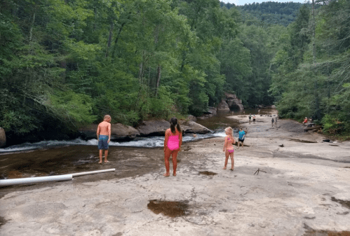 Children playing by a river in a wooded area, with people enjoying the water in the background.
