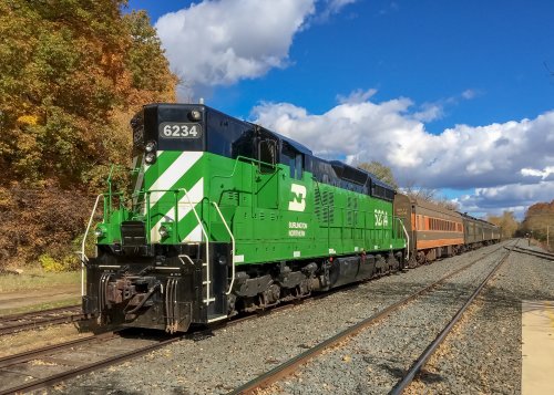 A green and black locomotive pulls a passenger train along a track surrounded by autumn foliage and a blue sky.