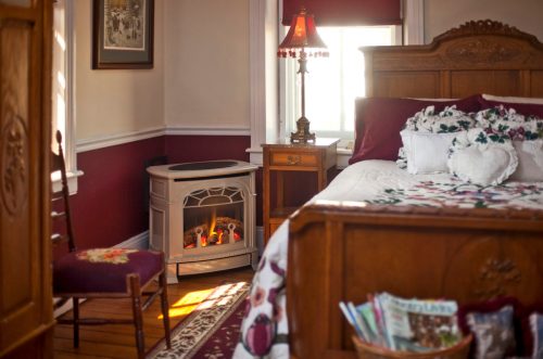 Cozy bedroom with a wooden bed, decorative pillows, a lamp, and a small electric fireplace in the corner.
