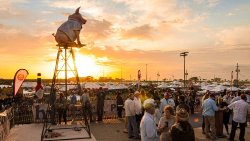 A pig statue in a blue shirt sits atop a tower at a lively outdoor event during sunset, with crowds and tents in the background.
