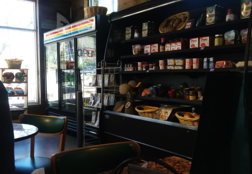 A cozy store interior featuring shelves of groceries, a glass fridge, and a table with chairs. Natural light from windows.