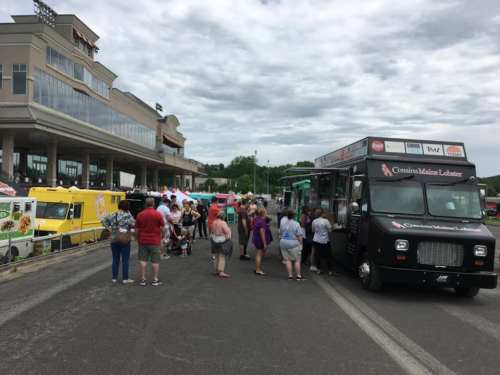 A crowd gathers around food trucks at an outdoor event, with a cloudy sky and a stadium in the background.