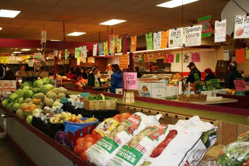 A bustling market scene with colorful produce displays, shoppers browsing, and vibrant signs hanging overhead.