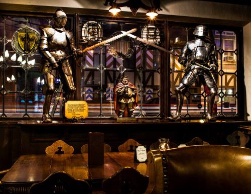 Two armored knights stand in a window display, flanking a statue, with a wooden table in the foreground of a dimly lit pub.