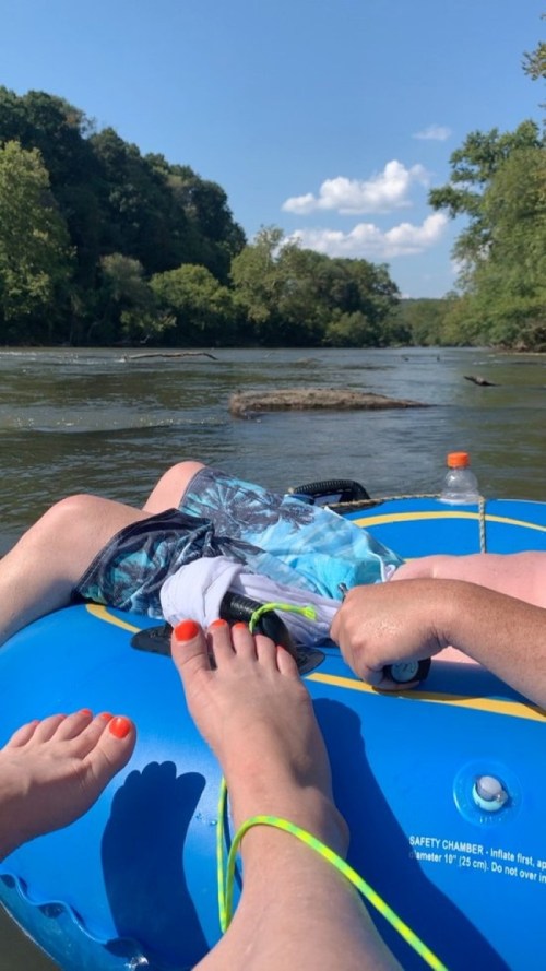 Two pairs of feet on a blue inner tube, floating on a river surrounded by trees and a clear blue sky.