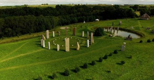 Aerial view of a stone circle surrounded by lush green fields and a small pond in the background.