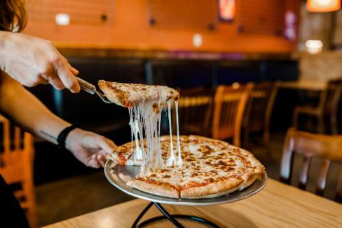A person lifts a slice of cheesy pizza from a round pizza on a serving tray in a restaurant setting.