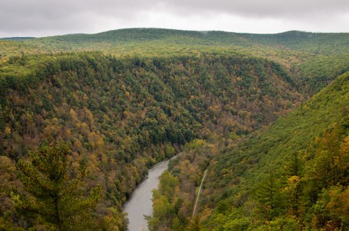 A scenic view of a winding river surrounded by lush, green hills and autumn-colored trees under a cloudy sky.