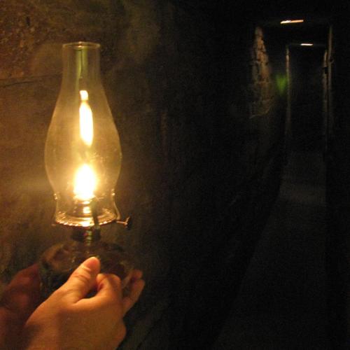 A hand holds a lit oil lamp in a dimly lit, narrow stone corridor.