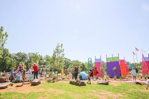 A vibrant playground with children playing, colorful structures, and trees under a clear blue sky.