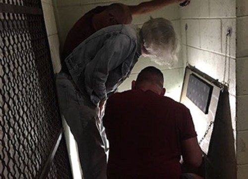 Two people lean over a small opening in a dimly lit room, examining something inside. A metal grate is visible.