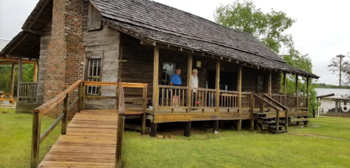 A rustic wooden house with a porch, surrounded by grass and trees, featuring two people standing on the porch.