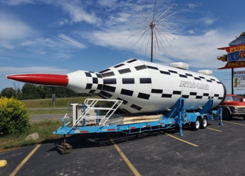 A large, checkered rocket-shaped structure on a trailer, parked outside a factory under a blue sky.