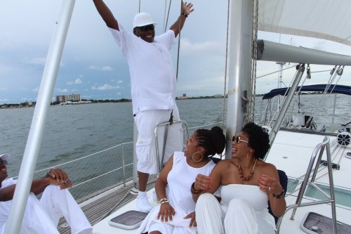 A group of friends in white outfits enjoying a sailboat ride, with one person joyfully standing on the deck.