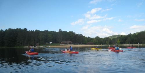 A group of kayakers paddles on a calm lake surrounded by trees and a distant cabin under a blue sky.