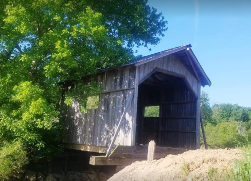 A rustic wooden covered bridge stands beside a tree, partially surrounded by dirt and greenery under a clear blue sky.