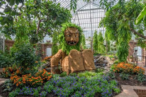 A large, moss-covered lion sculpture surrounded by vibrant flowers and lush greenery in a greenhouse setting.
