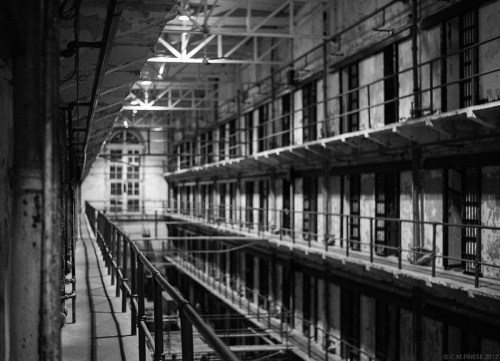 Black and white image of an empty prison corridor with barred cells and a high ceiling.