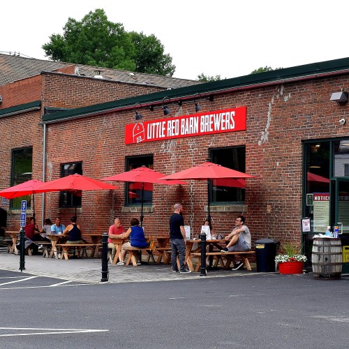 Exterior of Little Red Barn Brewers with outdoor seating under red umbrellas and patrons enjoying their time.