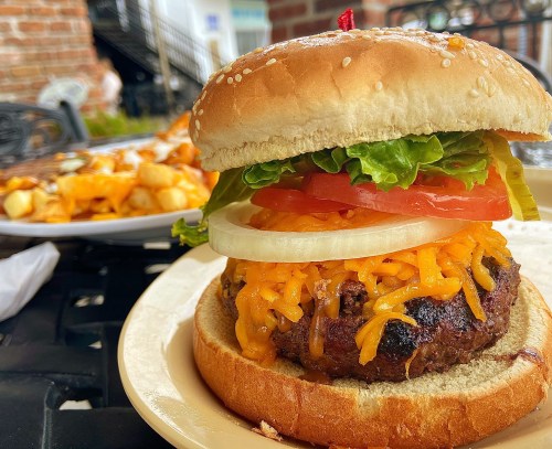 A close-up of a cheeseburger with lettuce, tomato, onion, and cheddar cheese on a sesame seed bun, with fries in the background.