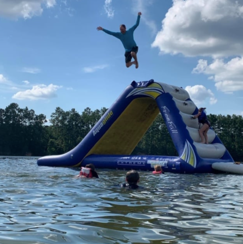 A person jumps off an inflatable water slide into a lake, while others swim nearby on a sunny day.
